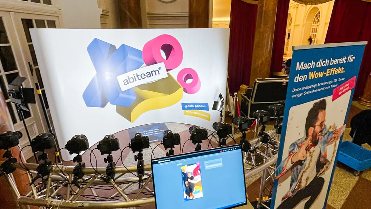 Bullet-Time-Booth-Setup im Festsaal: Halbkreis aus DSLR-Kameras auf Stativ, großes Backdrop mit abiteam*-3D-Logo, Monitor mit Live-Vorschau und Roll-up „Mach dich bereit für den Wow-Effekt."
