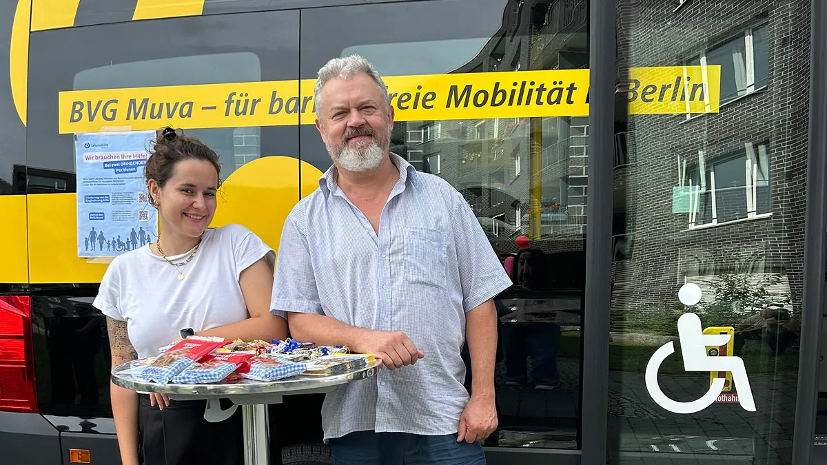 Denise als Event-Unterstützung mit BVG Muva Fahrer Witali neben einem BVG Muva Bus und dem Motto-Schild für barrierefreie Mobilität in Berlin. Daneben liegen Infomaterialien für Teilnehmende – Beispiel für zielgerichtete Kampagnen-PR und Event-Organisation.