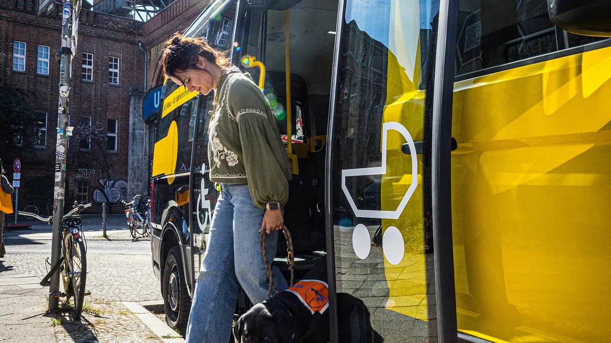 Eine Frau mit Blindenhund steigt problemlos aus dem BVG Muva Fahrzeug aus, vor Berliner Stadtkulisse. Kampagnenbild für barrierefreie Mobilität im Stadtalltag.